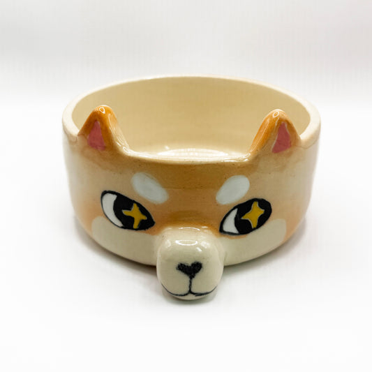 A ceramic bowl shaped like a Shiba Inu dog face, featuring a cream-colored body, tan and white facial markings, pink inner ears, and black and yellow starry eyes. The shiba inu is looking to the side giving it the iconic sussy look