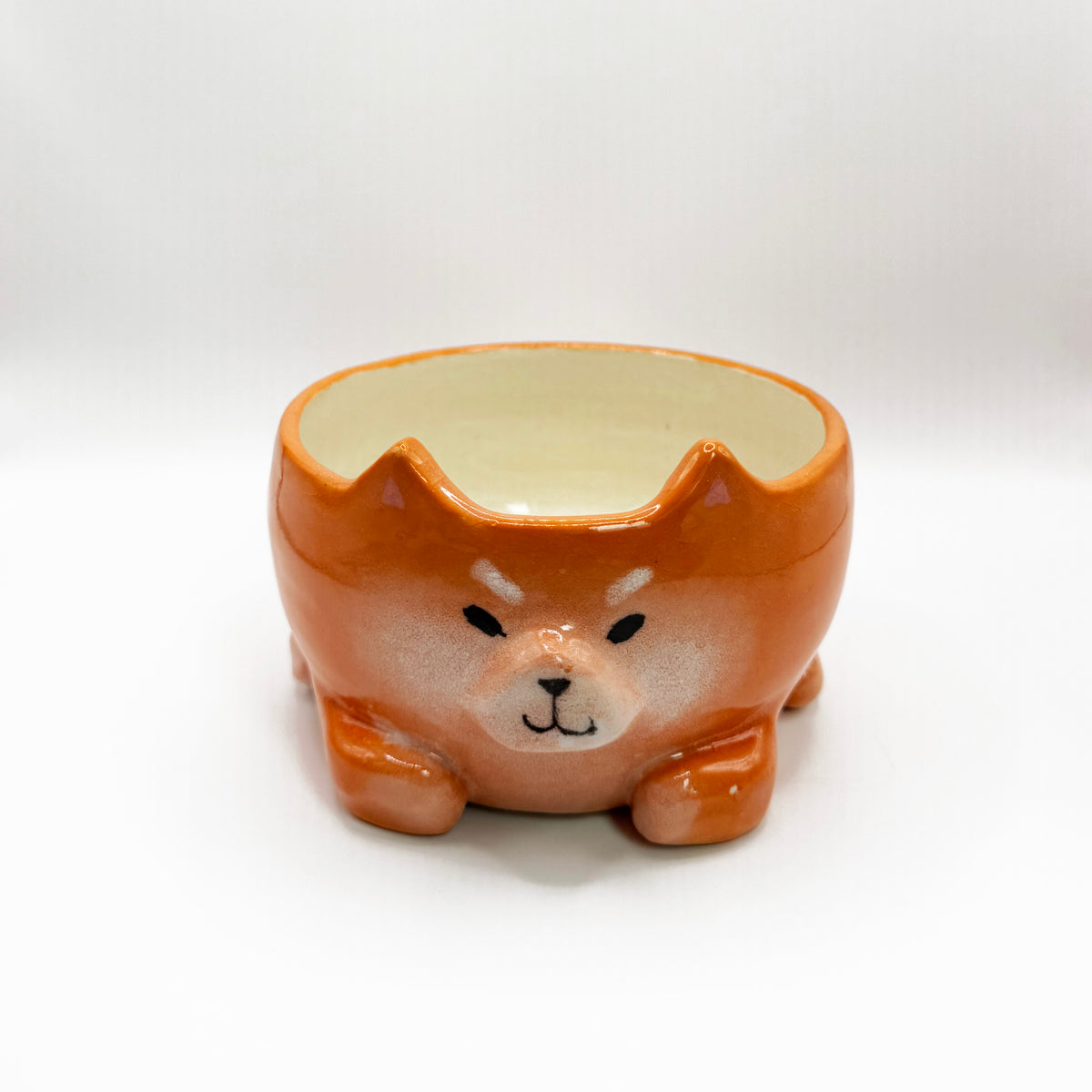 A small ceramic bowl shaped like a Shiba Inu dog head, featuring a glossy orange-brown glazed exterior and a cream-colored interior, set against a plain white background.
