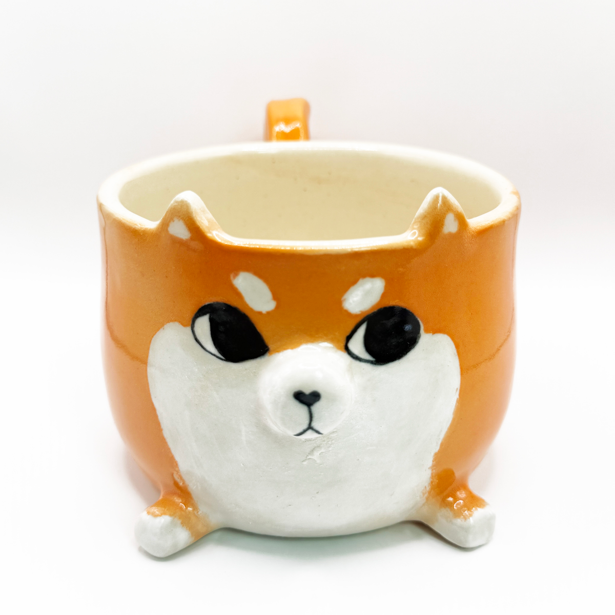 A ceramic coffee mug shaped like a Shiba Inu dog face, featuring a glossy orange and white glaze with black-painted eyes and nose, set against a plain white background. The shiba is looking to the side, giving the iconic side eye.