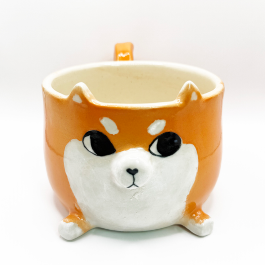 A ceramic coffee mug shaped like a Shiba Inu dog face, featuring a glossy orange and white glaze with black-painted eyes and nose, set against a plain white background. The shiba is looking to the side, giving the iconic side eye.