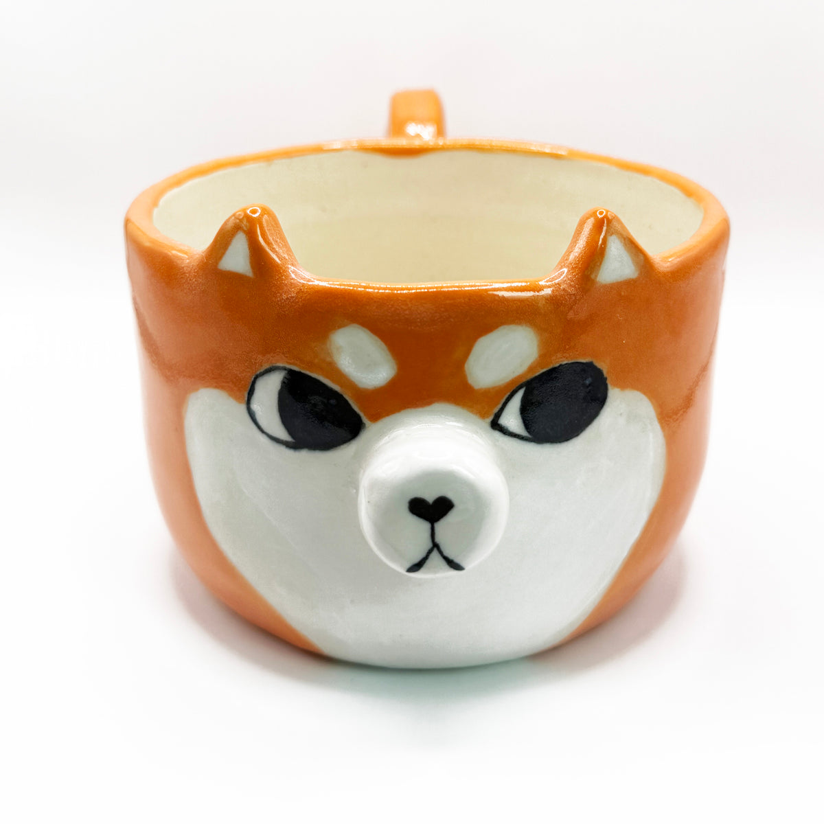 A ceramic coffee mug shaped like a Shiba Inu dog face, featuring an orange and white body with black eyes and nose details, set against a plain white background.The shiba inu is looking to the side giving it the iconic sussy look
