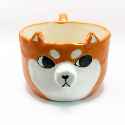 A ceramic coffee mug shaped like a Shiba Inu dog face, featuring an orange and white body with black eyes and nose details, set against a plain white background.The shiba inu is looking to the side giving it the iconic sussy look