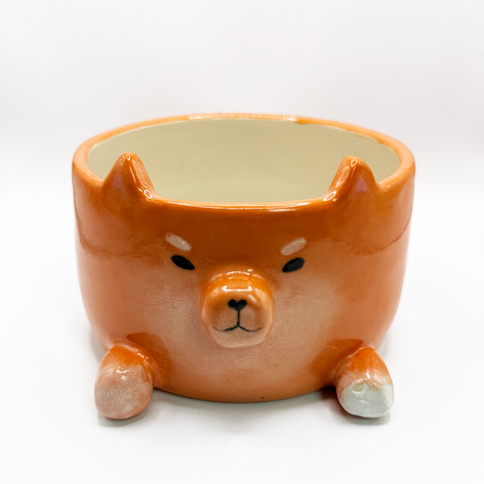An orange ceramic planter shaped like a Shiba Inu dog head, featuring a glossy finish, black painted eyes and nose, and small white accents on its paws and cheeks, set against a plain white background.