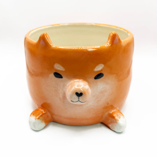 An orange ceramic planter shaped like a Shiba Inu dog head, featuring a glossy finish, black painted eyes and nose, and white accents on the paws and snoot, set against a plain white background. It has four legs