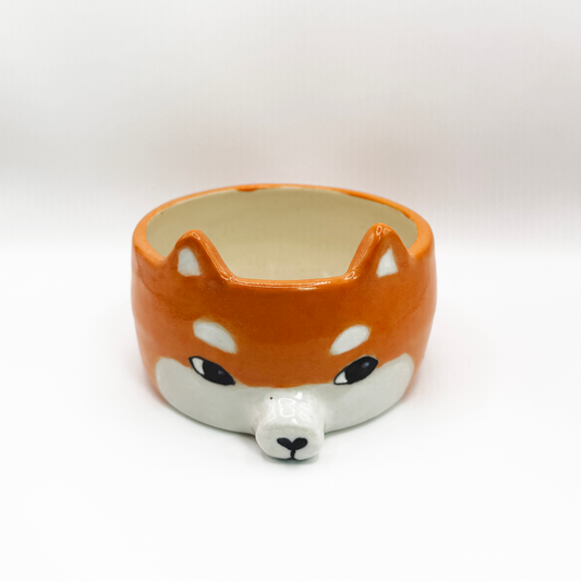 A ceramic bowl shaped like a Shiba Inu dog head, featuring a glossy orange and white glaze with black eyes and nose details, set against a plain white background. The shiba inu is looking to the side giving it the iconic sussy look