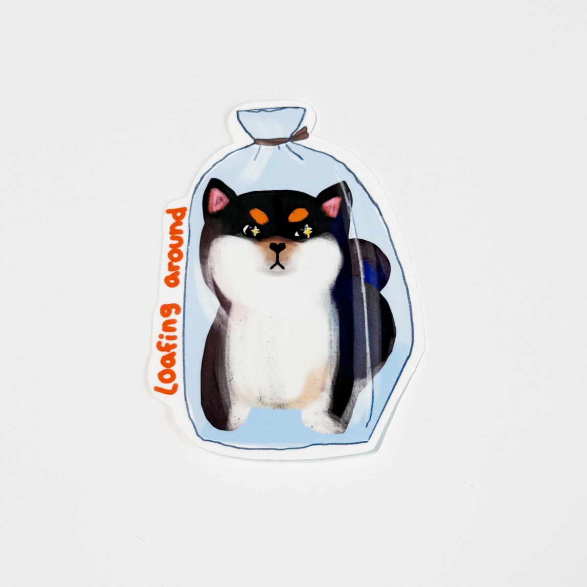 A die-cut sticker featuring an illustration of a black and white cat sitting inside a light blue plastic bag, with the phrase 'loafing around' written in orange text along the side, shown on a plain white background.