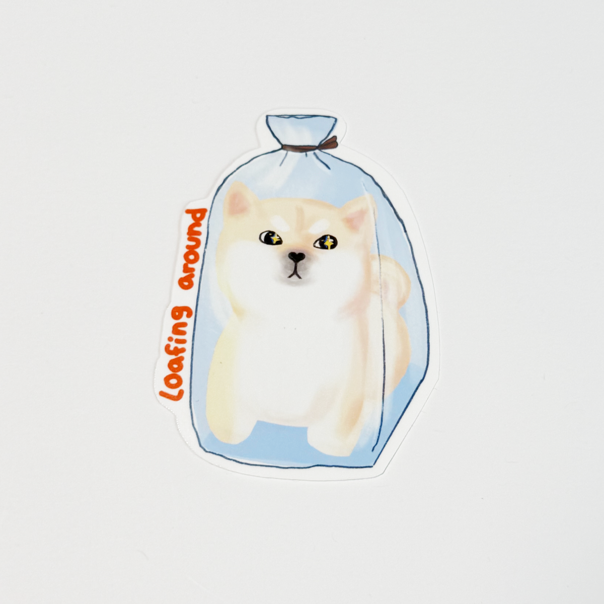 A die-cut sticker featuring a cute illustration of a cream-colored cat peeking out from inside a light blue drawstring bag, with the phrase 'loafing around' written in orange script on the side, shown on a plain white background.